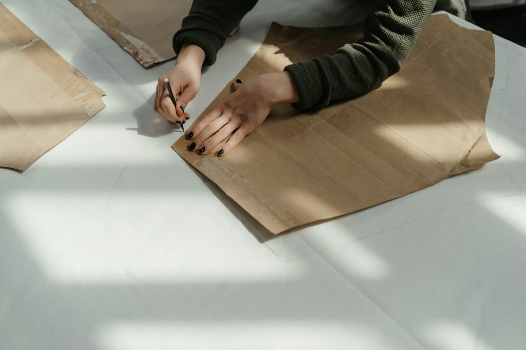 pexels-photo-4621569-4621569 Tailor tracing patterns on brown paper in a sunlit workshop, capturing the art of fashion design.