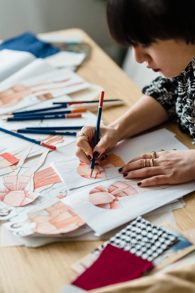 pexels-photo-7148060-7148060 From above of crop female designer drawing sketches while sitting at table with colorful fabrics and pencil in workshop